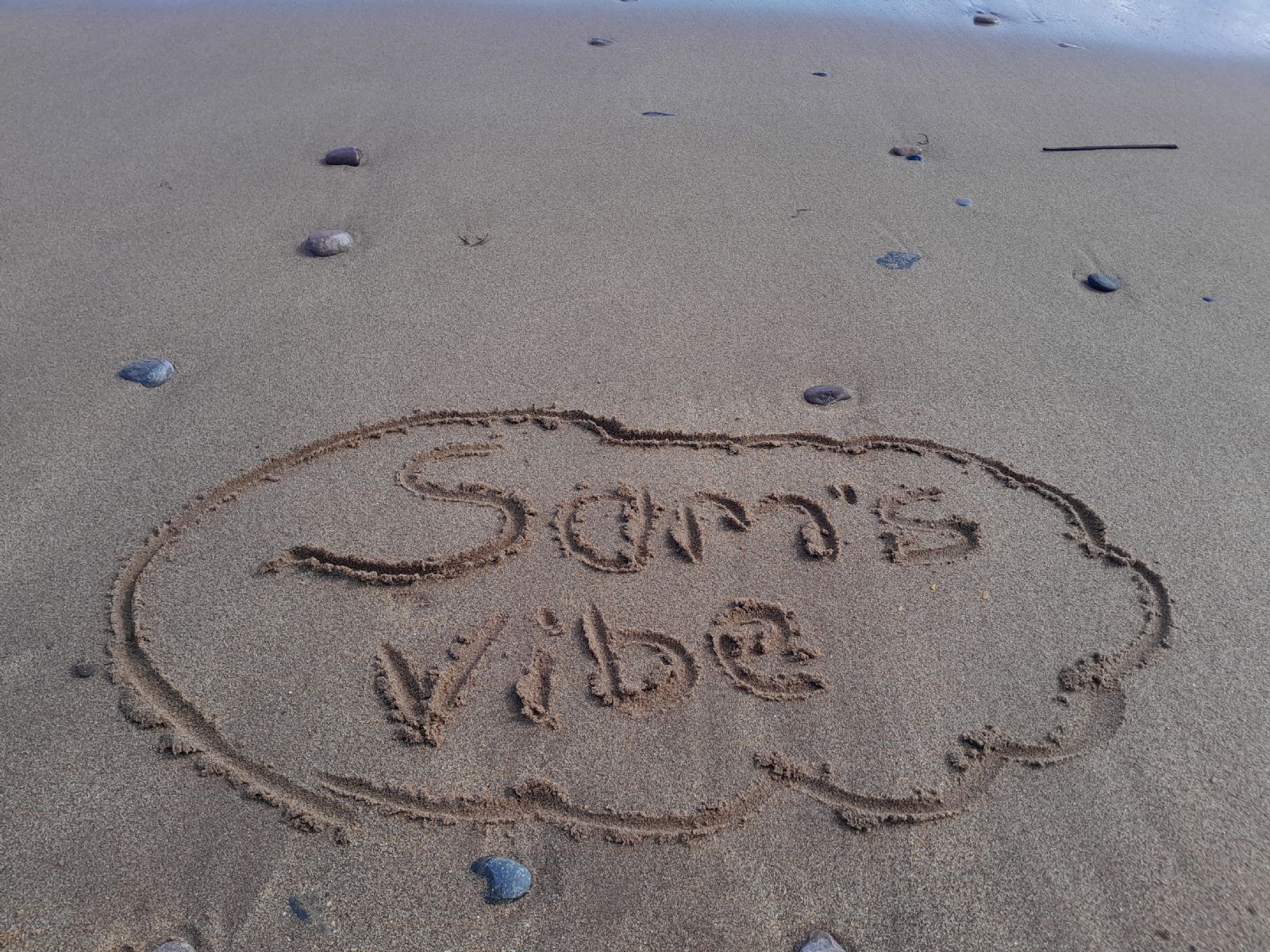 Picture of a beach with some pebbles. In the sand is words that state. Sam's Vibe